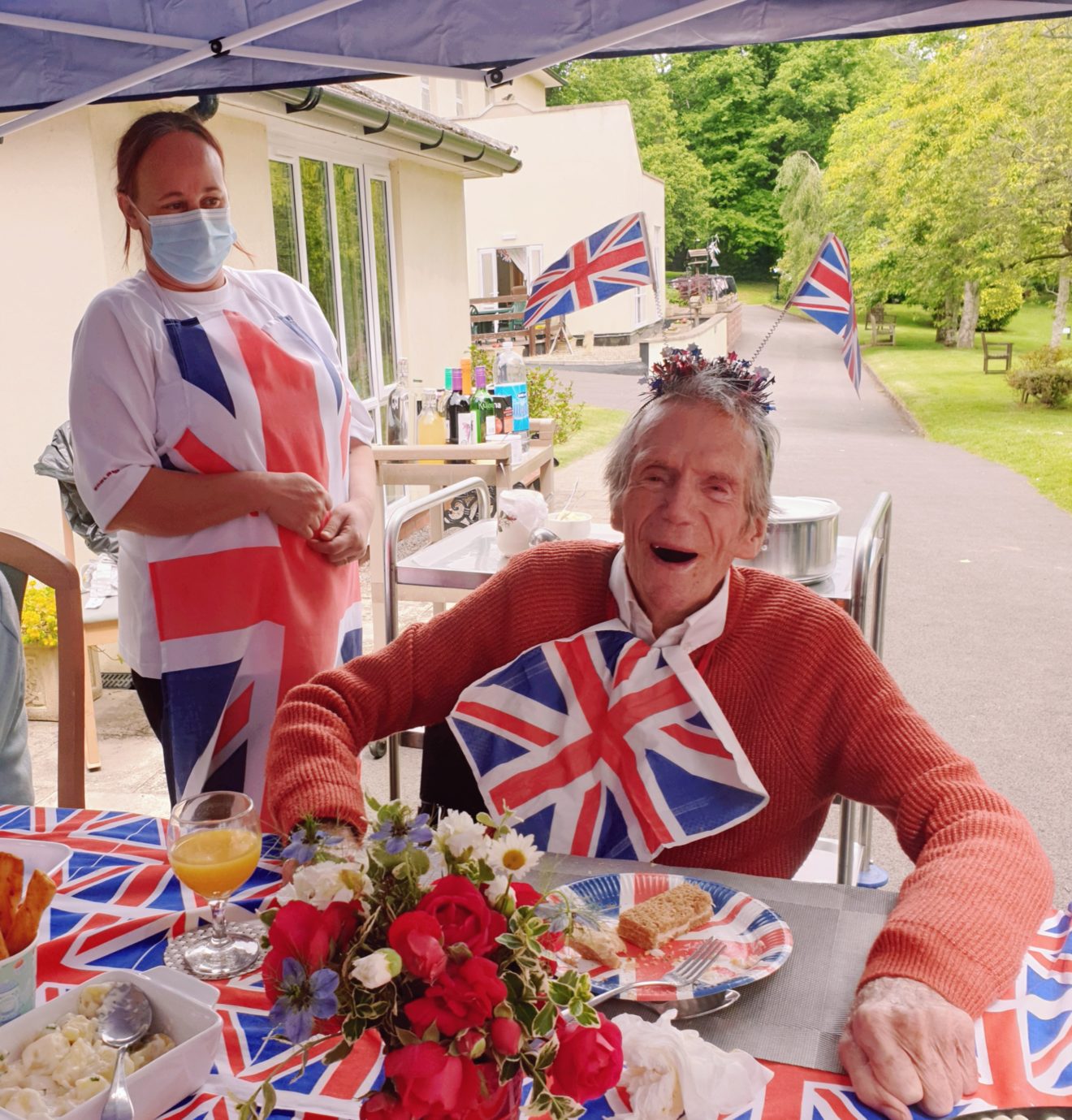 A man with a union jack napkin tucked into his collar and two union jacks in his hair looks very happy. A carer in a union jack apron and a mask looks on. 