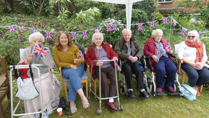 Six people seated outside in front of Union Jack bunting. Some of them are waving flags.