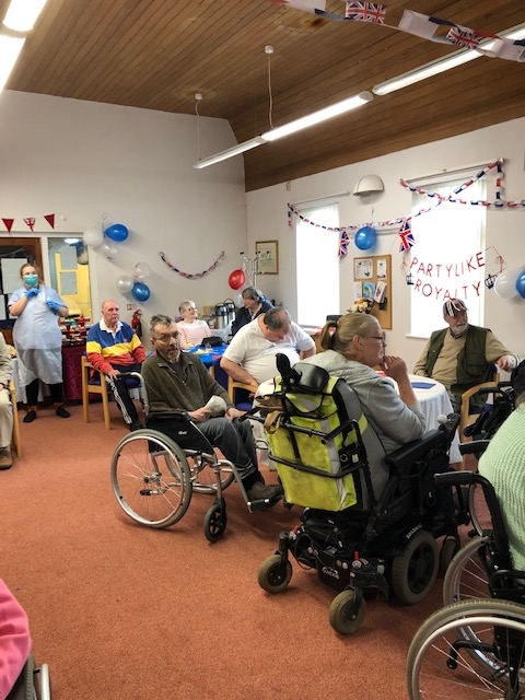 A group of people, some of wheelchairs, seated indoors. There is a banner in the background that reads 'Party Like Royalty'.