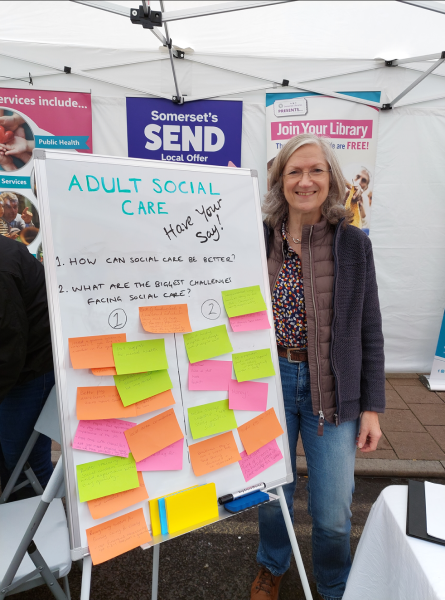 A woman standing in front of a white board which reads Adult Social Care: Have Your Say! Below there are post-it notes with responses.